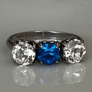 Op de afbeelding: Een zilveren ring met drie stenen. De middelste steen is een grote, blauwe saffier. De stenen aan beide kanten zijn kleiner en helder. De ring is gemarkeerd met "925 Sterling".
