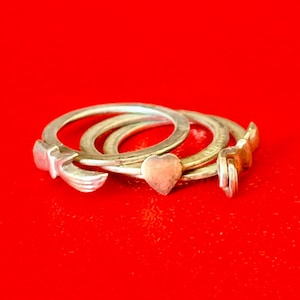 May include: Three silver rings are stacked on a red surface. One ring has a heart, the others have decorative elements. These rings are likely jewellery, suitable for everyday wear or special occasions. The rings are made of silver.