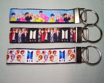 May include: Three keychains with different designs featuring the South Korean boy band BTS. Each keychain has a black and white ribbon with the band's logo and a silver key ring.