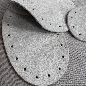 May include: Three grey leather patches with holes punched around the edges. The patches are oval shaped and are arranged on a grey textured surface.