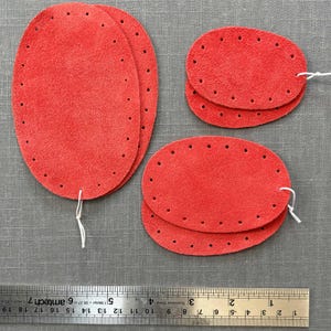 May include: Four red leather oval-shaped patches with holes around the edges. The patches are different sizes and are arranged on a grey fabric surface.