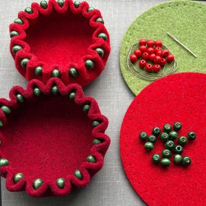 May include: Two red felt bowls with green bead trim. One bowl is empty, the other has green beads arranged in a star shape on a red felt circle. A green felt circle with red beads and a needle is in the background.
