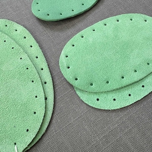 May include: A set of three green suede leather patches with stitching around the edges and holes for sewing. The patches are oval shaped and are stacked on top of each other.