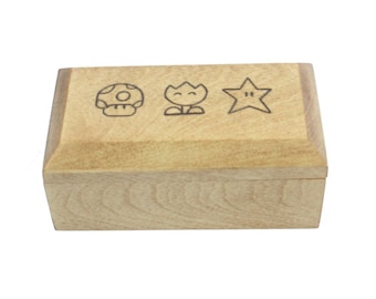 May include: A light wood box with a hinged lid. The lid is decorated with three black and white images of a mushroom, a flower, and a star.