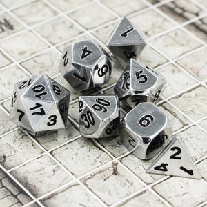 May include: A collection of seven silver polyhedral dice, ideal for role-playing games. The set includes a d4, d6, d8, d10, d12, d20, and a percentile die. Each die features black numbering on a metallic silver surface. The dice are displayed on a stone-effect surface.