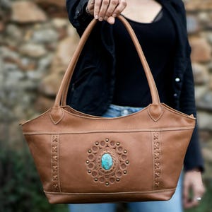 Puede incluir: Un bolso tote de cuero marrón con una pieza central de piedra turquesa y costuras decorativas. El bolso tiene un asa y el texto "SIBQ YANKE" y "Exclusive Desings with all magic of the GEMSTONES".