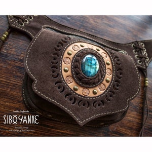 Puede incluir: Bolsa de cinturón de cuero marrón con intrincados recortes y una gran piedra preciosa azul en el centro. La bolsa tiene una cremallera y un detalle decorativo de encaje.
