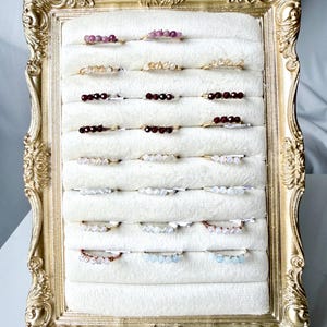 May include: A collection of twenty-four delicate wire-wrapped rings featuring small gemstone beads in various colors, including pink, yellow, deep red, white, and light blue, displayed in rows on a white velvet insert within a gold ornate frame.