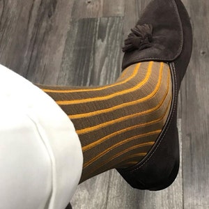 Puede incluir: Un par de mocasines de ante marrón con un detalle de borla. Los mocasines se llevan con calcetines a rayas marrones y amarillas.