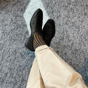 Peut inclure: Une paire de mocassins en daim noir avec un d&eacute;tail de boucle dor&eacute;e. Les mocassins sont port&eacute;s avec des chaussettes ray&eacute;es et un pantalon beige.