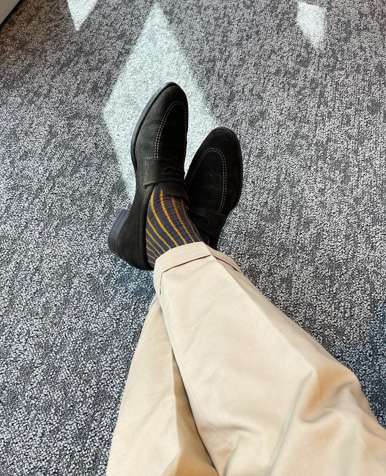 Peut inclure: Une paire de mocassins en daim noir avec un d&eacute;tail de boucle dor&eacute;e. Les mocassins sont port&eacute;s avec des chaussettes ray&eacute;es et un pantalon beige.