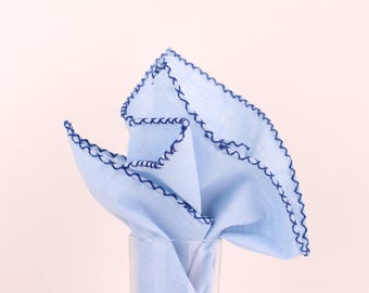 May include: A light blue handkerchief with a dark blue scalloped edge, partially displayed in a clear glass. The fabric is folded and arranged to show the decorative trim. The background is a soft, neutral color.