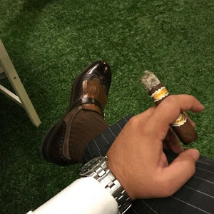 Peut inclure: Une personne portant une chemise blanche, une chaussure en cuir marron, une montre argentée et un pantalon noir à rayures blanches tient un cigare brun avec un bandeau doré.
