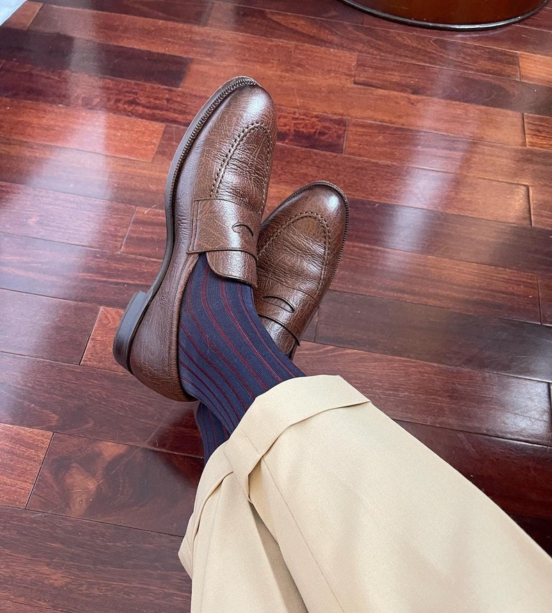 Peut inclure: Mocassins en cuir marron avec une bride &agrave; penny. Les mocassins sont port&eacute;s avec des chaussettes ray&eacute;es bleues et rouges et un pantalon beige.