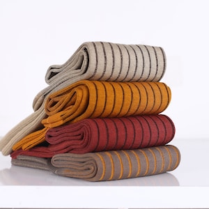 Puede incluir: Una pila de cuatro pares de calcetines doblados con un patrón de rayas verticales. Los calcetines son de colores beige, amarillo mostaza, rojo oscuro y marrón con rayas en contraste, dispuestos en un montón ordenado sobre un fondo blanco.