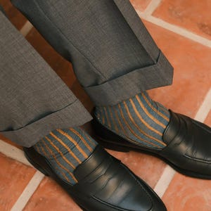 May include: A pair of black leather penny loafers worn with blue and orange striped socks and grey cuffed trousers. The shoes feature a classic slip-on design with a horizontal strap across the vamp and a rounded toe, paired with textured socks.