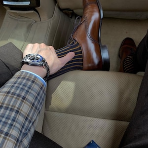 Peut inclure: Un homme portant une veste de costume &agrave; carreaux, un pantalon marron et une chaussette ray&eacute;e bleu et jaune. Il porte une chaussure en cuir marron Oxford et une montre argent&eacute;e avec un cadran bleu. Il est assis dans une voiture avec un si&egrave;ge en cuir beige.