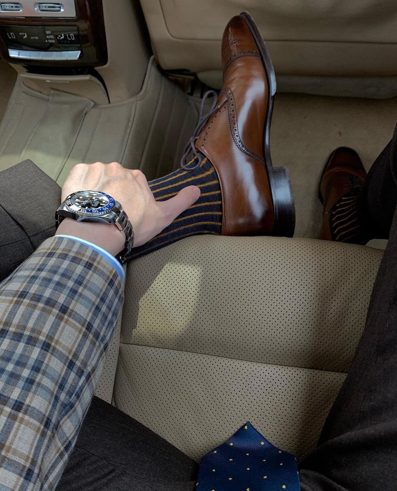 Peut inclure: Un homme portant une veste de costume &agrave; carreaux, un pantalon marron et une chaussette ray&eacute;e bleu et jaune. Il porte une chaussure en cuir marron Oxford et une montre argent&eacute;e avec un cadran bleu. Il est assis dans une voiture avec un si&egrave;ge en cuir beige.