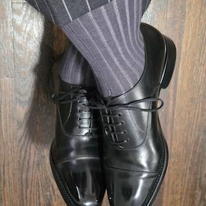 Puede incluir: Un par de zapatos oxford de cuero negro con acabado pulido y cordones negros finos, usados con calcetines grises acanalados y pantalones gris oscuro a rayas. Los zapatos presentan un diseño clásico con puntera y costuras visibles.
