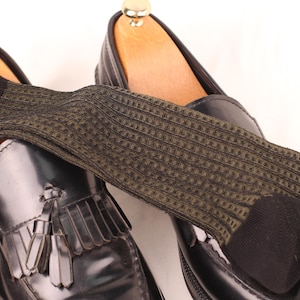 May include: A pair of black leather loafers with tassels, paired with olive green patterned socks. The socks have a textured design and are draped over the shoes, showcasing a classic style.