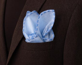 May include: A light blue pocket square with a stitched border is displayed in the breast pocket of a dark brown pinstripe blazer. The pocket square is folded to show its edges. The blazer is worn over a black turtleneck.