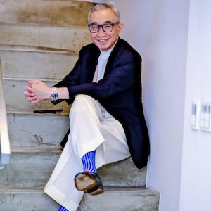 May include: A dark navy blue blazer worn over a white mock neck shirt, paired with white trousers, blue and white vertical striped socks, and black loafers with red accents. A silver wristwatch is worn on the left wrist and dark-rimmed glasses.