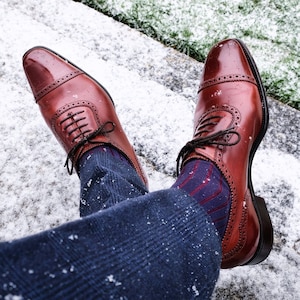 Puede incluir: Un par de zapatos Oxford de cuero marrón con detalles de brogue. Los zapatos se llevan con calcetines azules estampados y pantalones azules. Los zapatos están sobre una superficie nevada.