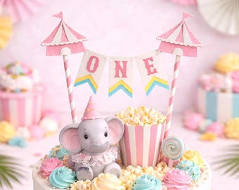 May include: A whimsical, pastel-colored cake decorated for a first birthday. The cake features pink and white stripes, colorful frosting, and a charming elephant figurine. A banner spells out "ONE" above the cake, with popcorn and a lollipop.
