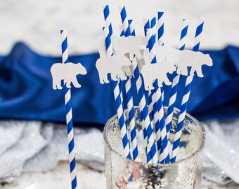 May include: A glass jar filled with blue and white striped paper straws, each topped with a white polar bear cutout. The straws are arranged in a cluster, with some leaning against the side of the jar. The jar is sitting on a white surface, with a blue satin fabric draped behind it.
