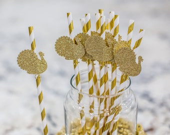 May include: A glass jar filled with gold and white striped paper straws with gold glitter turkey cutouts attached to the tops. The jar is sitting on a white surface with gold glitter.
