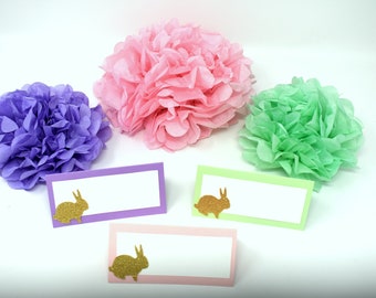 May include: Four place cards with gold glitter bunny silhouettes on purple, pink, and green cardstock. The place cards are arranged in a row on a white surface. There are three tissue paper pom-poms in the background, one purple, one pink, and one green.