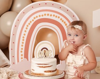 May include: A baby girl sits in front of a cake decorated with a rainbow and the word "ONE". The cake is on a white cake stand. The background is decorated with balloons, a teepee, and a large number one.