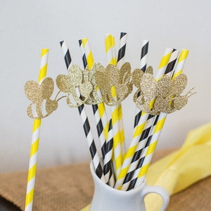 May include: A collection of decorative paper straws in a white pitcher. The straws feature yellow and white stripes, black and white stripes, and are adorned with gold glitter bee cutouts. A yellow cloth is in the background.