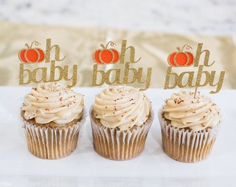 May include: Three cupcakes with brown frosting and gold glitter toppers that say "Oh Baby" with a pumpkin design.