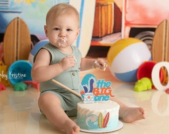May include: A baby in a green onesie sits in front of a beach-themed backdrop with a surfboard and a van. The baby is eating cake with frosting on their face. The cake is decorated with a blue and white surfboard and the words "The Big One".