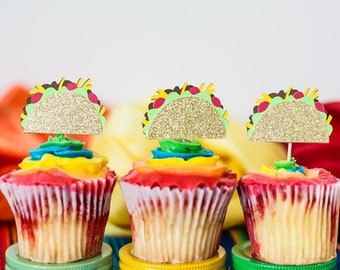 May include: Three colorful cupcakes decorated with rainbow frosting and topped with gold glitter taco cupcake toppers. The cupcakes are sitting on a colorful Mexican blanket.