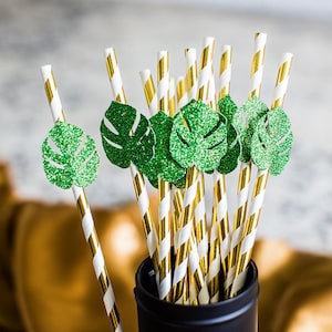 May include: A bunch of gold and white striped paper straws with green glitter monstera leaf cutouts attached to the top. The straws are in a black container.