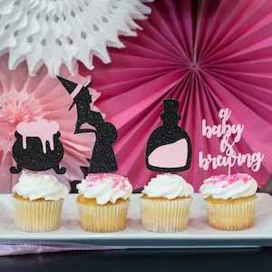 May include: Four cupcakes decorated with black and pink glitter toppers. The toppers feature a pregnant witch, a cauldron, a bottle with a pink potion, and the text "a baby is brewing".