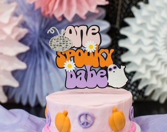 May include: A pink cake with orange and purple decorations. The cake is topped with a cake topper that says "one spooky babe" in a retro font. The cake topper also features a disco ball, a ghost, and daisies.