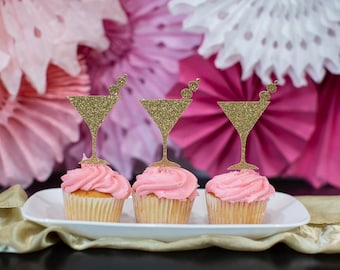 May include: Three pink cupcakes with gold glitter martini glass toppers. The cupcakes are on a white plate and a gold tablecloth.