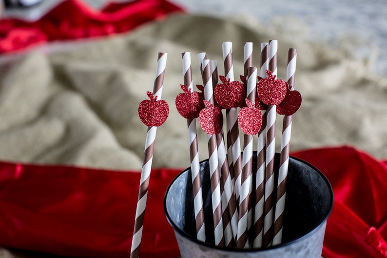 May include: A close-up of a group of brown and white striped paper straws with red glitter apple decorations. The straws are in a silver metal bucket.