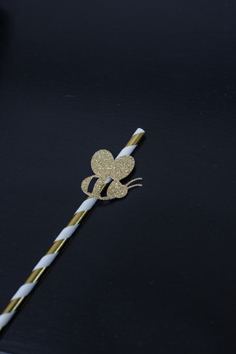 May include: A white and gold striped paper straw with a gold glitter bee cutout attached to the top.