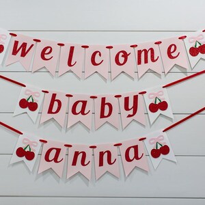 May include: A three-tiered banner with the words "welcome baby anna" in red script on pink pennants. Each pennant is adorned with a red cherry illustration. The banner is strung with red ribbon against a white wood-paneled background.