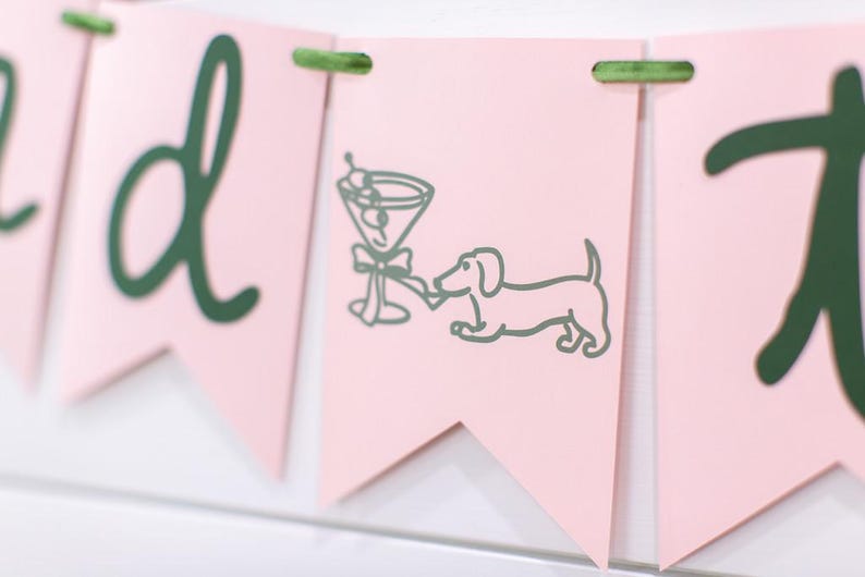 May include: Pink banner with green text and accents. One banner features a martini glass and a dachshund dog illustration. The banner is strung together with green twine. The overall theme is party or celebration.