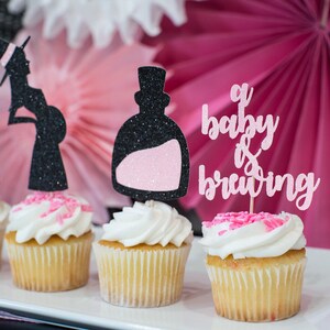 May include: Four cupcakes decorated with black and pink glitter toppers. The toppers feature a witch, a potion bottle, and the text "a baby is brewing".