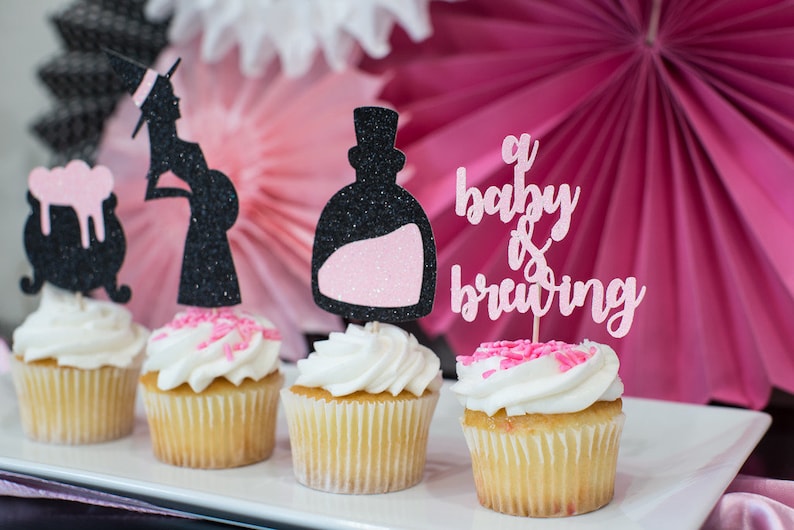 May include: Four cupcakes decorated with black and pink glitter toppers. The toppers feature a witch, a potion bottle, and the text "a baby is brewing".