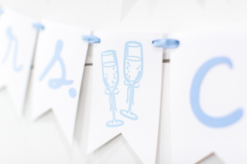 Op de afbeelding: Een witte wimpelslinger met lichtblauwe letters en een centrale afbeelding van twee champagneglazen. De afzonderlijke wimpels zijn verbonden door een lichtblauw lint dat door gaten aan de bovenkant van elk driehoekig papieren stuk is geregen.