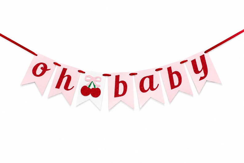 May include: A decorative banner featuring light pink pennants strung on a red cord. The letters spell out "oh baby" in a glittery red font. A white pennant in the center displays two red cherries with a small light pink bow above them.