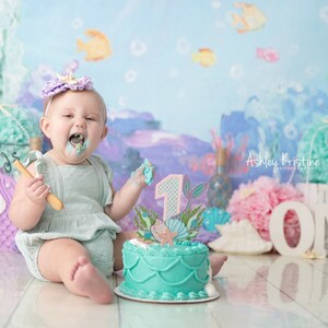 May include: A baby girl wearing a light blue dress and a purple flower crown is sitting in front of a turquoise and purple cake decorated with a mermaid tail and a number one. The cake is on a white plate and the baby is holding a wooden spoon. The background is a blue and purple ocean-themed backdrop with the words "Mermaids" and "ONE" visible.