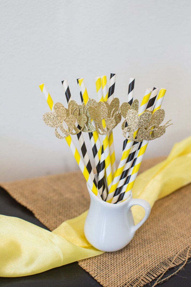 May include: A collection of decorative paper straws in a white pitcher. The straws have yellow and black stripes, with gold glitter butterfly accents. The pitcher sits on a burlap and yellow fabric background.